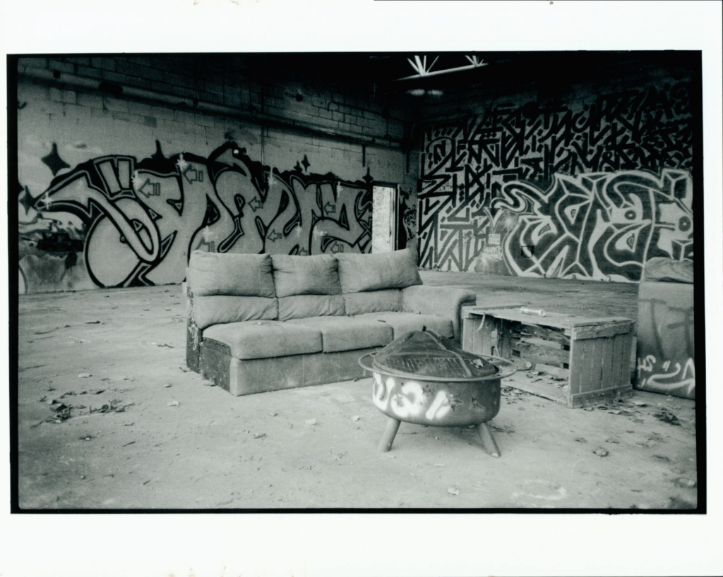 Black and white photo of a couch and a table in a room with graffiti on the walls, debris and firepit on the floor